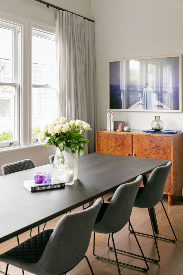 A bright, refined dining room with a large dark rectangular dining table surrounded by upholstered charcoal dining chairs. Tall white-trimmed windows with full-length light curtains bring in abundant daylight. A warm mid-century-style wooden sideboard sits against the wall beneath framed artwork, with minimal decorative objects on top. Light timber flooring and neutral walls complete the calm contemporary interior.
