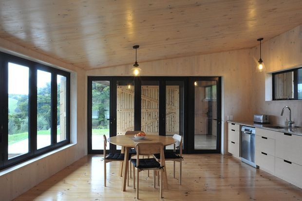 A warm, compact open-plan kitchen and dining area lined with natural timber on the floor, walls, and ceiling. A light timber dining table with chairs sits centrally beside large black-framed windows and glazed doors. The kitchen has pale minimalist cabinetry, a dark benchtop, integrated appliances, and a stainless-steel sink. Pendant lights and views to greenery create a bright, calm connection to the outdoors.