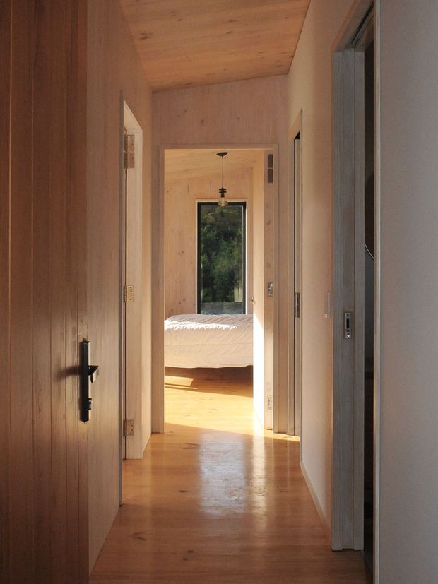 A warmly lit narrow hallway lined with natural timber wall and ceiling panels, leading through an open doorway into a bright bedroom. The corridor has polished timber flooring, simple flush doors, dark hardware, and a minimal pendant light visible in the bedroom. Sunlight enters through the bedroom window, creating a calm, cohesive timber-lined interior.