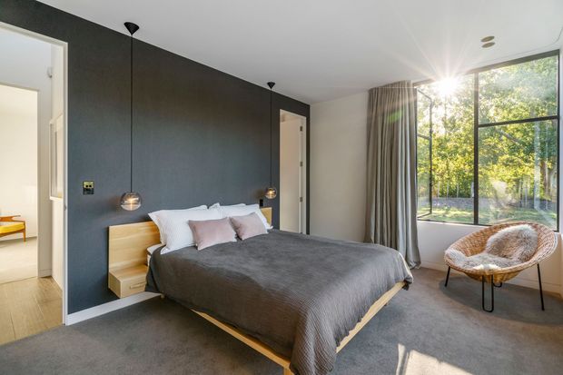 A contemporary bedroom with a low timber platform bed, dark charcoal feature wall, integrated timber bedside shelves, and two suspended pendant lights. The room has soft grey carpet, neutral curtains, white bedding with pale blush cushions, and a large black-framed window overlooking leafy greenery. A woven lounge chair sits near the window, and open doorways lead to adjacent spaces.