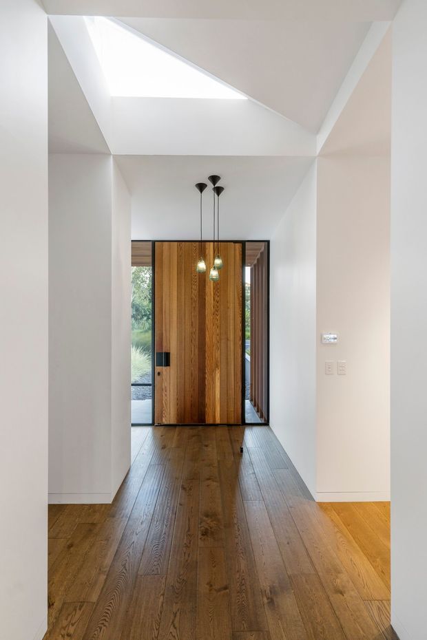 A minimalist residential entry hall with a tall timber pivot front door framed by dark glazing and sidelights. Warm timber flooring extends through the corridor, contrasting with white walls and a sculptural angled ceiling containing a large skylight. Three slim black pendant lights hang above the entry, with garden greenery visible beyond the glazed doorway.