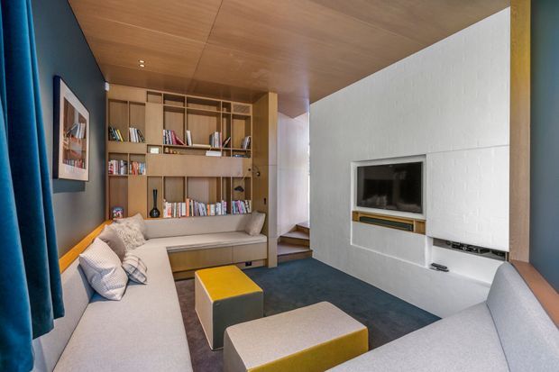 Compact contemporary media room with a large L-shaped upholstered sectional sofa, yellow and grey ottomans, built-in timber shelving filled with books, and a wall-mounted television set into a white textured feature wall. The room has a warm timber ceiling, dark blue walls and carpeted flooring, with stairs leading beyond the shelving area.