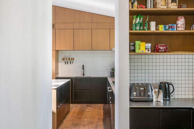 A compact, galley-style kitchen viewed through a doorway, with warm timber upper cabinetry, dark lower cabinetry, open timber pantry shelving, white grid-tile splashbacks, stainless-steel benchtops, and a parquet timber floor. A toaster and kettle sit on the foreground counter, while the main kitchen includes a sink, cooktop area, and wall-mounted utensils.
