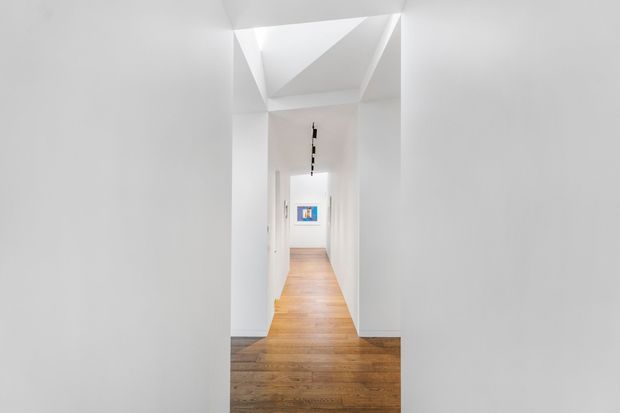 A bright minimalist interior hallway with white walls and sculptural angular ceiling planes. Warm timber flooring extends through the corridor, leading toward a small framed artwork, while a slim black linear light fixture runs along the ceiling.