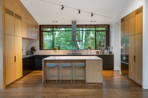 A spacious contemporary kitchen with a central island and three stools, extensive warm timber cabinetry, dark lower cabinets, and a light benchtop. A long horizontal window provides abundant natural light and views of leafy greenery. Integrated appliances and a slimline rangehood contribute to the clean, streamlined design, while timber flooring adds warmth.