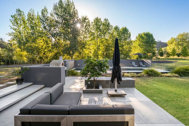 A landscaped outdoor entertaining area with a contemporary built-in charcoal-grey sectional lounge, pale tiled paving, raised planters, and a closed black parasol. A low outdoor fireplace or grill structure and additional seating areas sit beyond, surrounded by mature trees and lawn in warm late-afternoon sunlight.