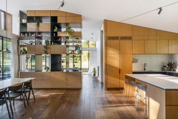 A bright, open-plan contemporary kitchen and dining area with extensive warm timber cabinetry and joinery. A large, partially open shelving and storage partition separates the dining space from the kitchen while maintaining visual connection. The kitchen includes a white island benchtop, integrated timber cabinetry, a small square-tile splashback, bar seating, and black plumbing fixtures. Full-height glazing and timber flooring create an airy, warm interior, with track lighting and indoor plants adding detail.