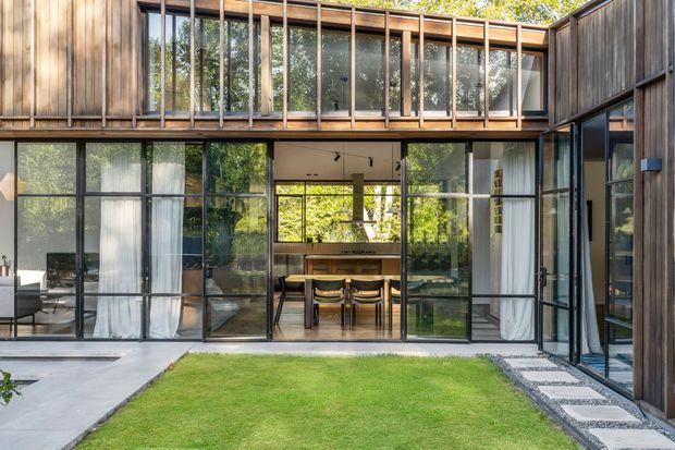 A contemporary timber-clad two-storey courtyard house with extensive black-framed floor-to-ceiling glazing opening onto a compact lawn. The central glazed opening reveals a dining area with a timber table and chairs, while upper-level glazing and timber screening create a private, light-filled composition around the courtyard. Concrete paving and stepping stones border the grass.
