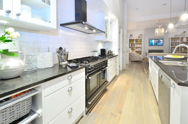Bright, narrow galley kitchen with white shaker-style cabinetry, dark stone benchtops, a stainless steel range cooker and overhead extractor, white subway-tile splashback, and light timber flooring. The kitchen opens toward a living area with built-in shelving and pendant lighting.