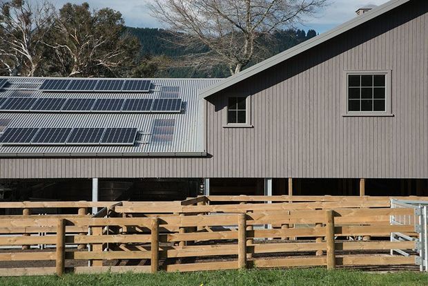 Exterior view of a raised rural barn or agricultural building with dark vertical cladding, a corrugated metal pitched roof, and extensive photovoltaic solar panels. Timber livestock fencing and an elevated open undercroft are visible in the foreground, with wooded hills behind.