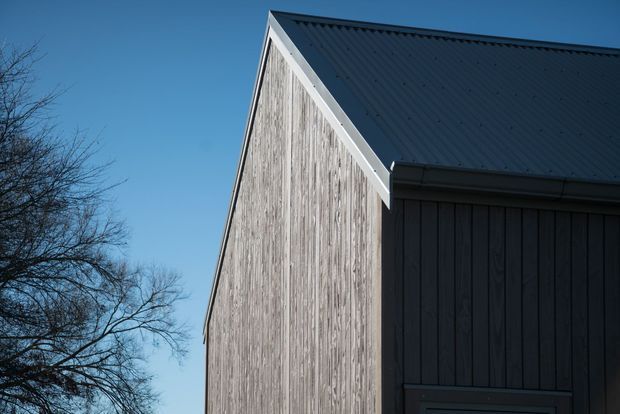 Close-up exterior view of a simple gabled building, featuring weathered vertical timber cladding on the front gable and darker vertical wall cladding on the side. The roof is a dark corrugated metal sheet with light contrasting fascia and guttering. Bare tree branches and clear blue sky frame the structure.