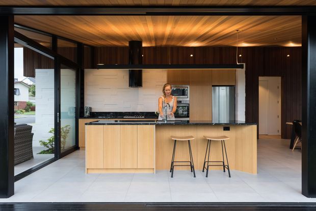 A contemporary open-plan kitchen viewed from a large glazed opening. The room features extensive light timber cabinetry and a matching island with two stools, a dark stone or composite benchtop, integrated appliances, a white textured splashback, pale tiled flooring, and a warm timber-lined ceiling. Black-framed floor-to-ceiling glazing opens to an outdoor area, while dark structural framing and pendant lighting add contrast.