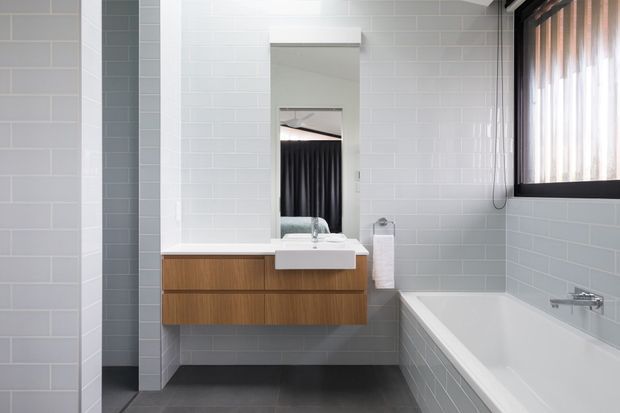 Bright contemporary bathroom with white subway-tiled walls, a floating timber vanity with white basin, wall mirror, freestanding-style built-in bathtub, dark grey tiled floor, and a black-framed window with sheer curtains. The space has clean lines and a restrained neutral palette with warm wood joinery.