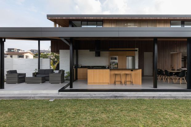 A contemporary open-plan indoor-outdoor living area viewed from the lawn, with a covered patio, outdoor seating, and a kitchen island opening through large glazed doors. The house features strong horizontal lines, dark metal framing, warm vertical timber cladding, timber ceilings, and a flat roof. A dining area is visible to the right, while neighbouring homes and landscaping appear in the background.