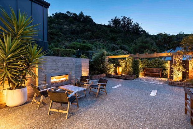 A landscaped outdoor entertaining terrace at dusk, featuring a built-in stone fireplace, outdoor seating and dining furniture, large planters, timber screening, and integrated warm lighting. Dense hillside vegetation forms the backdrop, while a dark-clad building edge is visible at left.