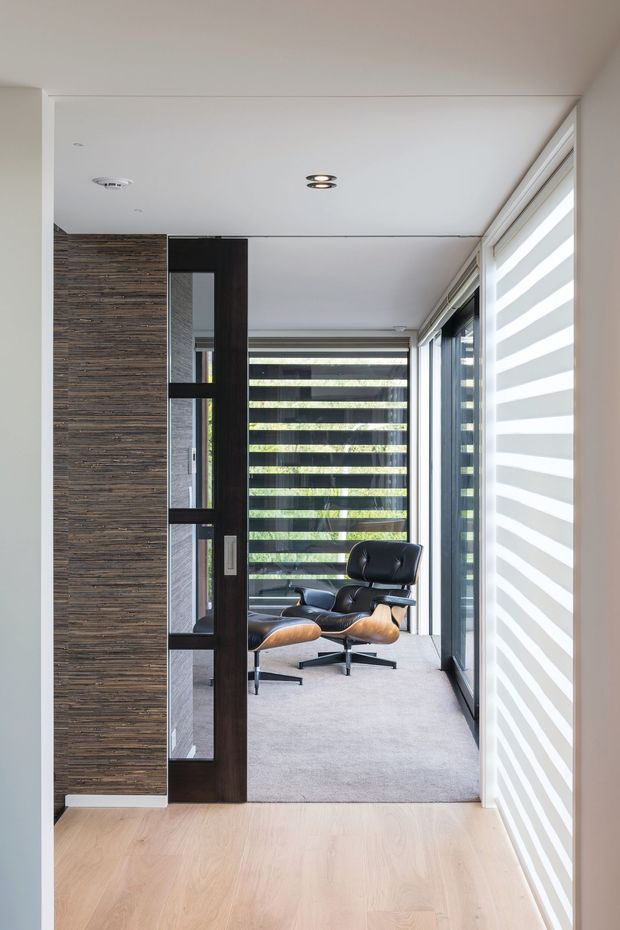 A narrow, modern home office or study viewed through a partial partition. The room features a dark timber-framed glazed divider, textured horizontal timber wall cladding, floor-to-ceiling glazing with striped roller blinds, a black leather lounge chair with matching ottoman, recessed ceiling lights, and light grey carpet. The foreground has pale timber flooring and the space is bright and restrained.