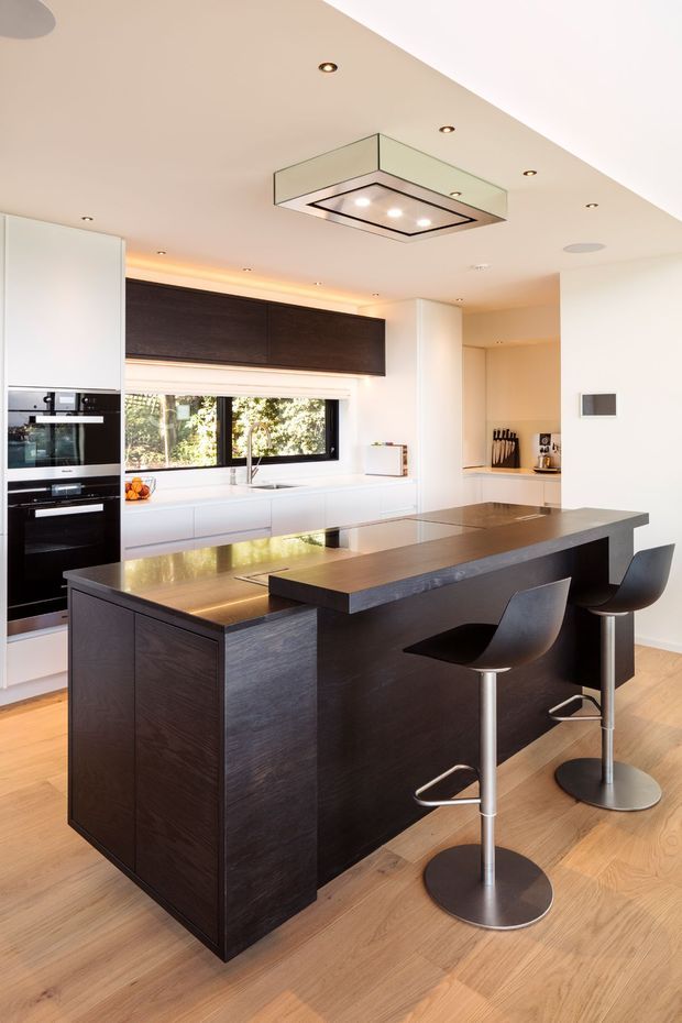 A bright contemporary kitchen featuring a large dark timber island with an integrated raised dining counter and two modern bar stools. White full-height cabinetry, built-in black appliances, a dark overhead cabinet run, light timber flooring, a horizontal window, and a prominent glass-and-metal rangehood create a refined high-contrast interior.