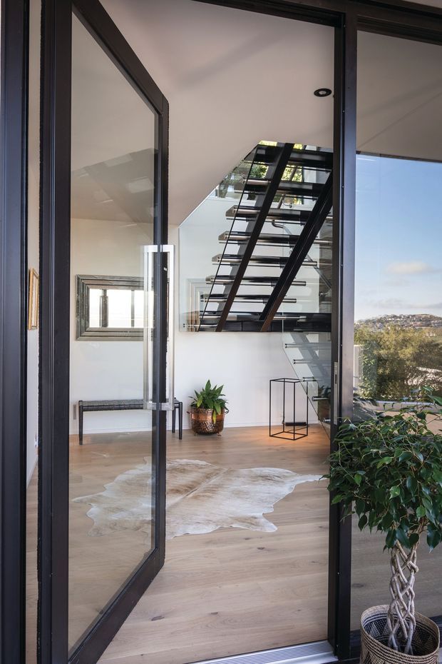 A bright contemporary interior viewed through an open black-framed glazed door, featuring a floating staircase with dark metal stringers and treads, pale timber flooring, white walls, a skylight or glazed roof opening, and a sculptural potted plant. A distant residential landscape is visible beyond the glazing.