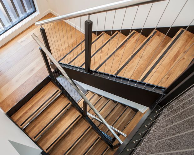 A contemporary multi-level stairway viewed from above, featuring warm timber stair treads and flooring contrasted with black-painted structural stringers, balustrade posts, and slim vertical cable infill. A polished metal handrail follows the stair edges, with light walls and a window visible alongside the landing.