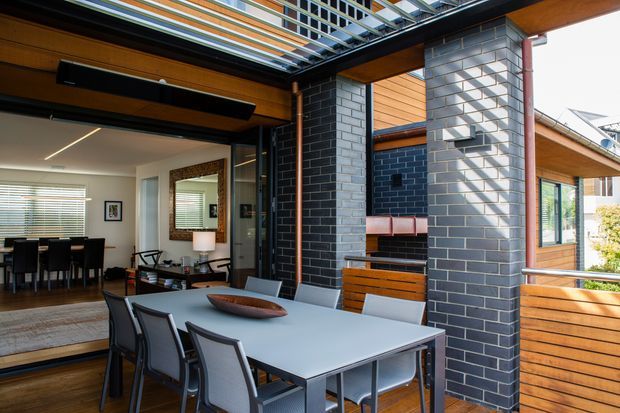A covered outdoor dining terrace adjoining a contemporary home, with a large rectangular dining table and chairs, timber decking, dark brick feature walls, timber-lined soffits, and a built-in outdoor fireplace or barbecue niche. A glass-and-metal pergola roof provides filtered sunlight. The adjoining interior dining and living areas are visible through wide openings.
