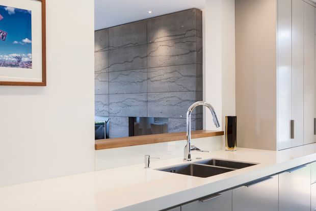 Contemporary kitchen interior focused on a bright white island benchtop with an undermount double sink and tall curved chrome tap. Full-height white cabinetry lines the right side, while a timber-framed opening or shelf and a large mirror/reflection reveal a textured grey concrete feature wall beyond. A framed artwork is partially visible on the left. The space has clean lines, integrated joinery, recessed lighting, and a restrained neutral palette with timber and chrome accents.