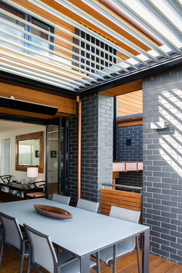 A contemporary outdoor dining courtyard or covered terrace adjoining an interior living area. The space features a large pale dining table with modern chairs, warm timber decking and wall cladding, dark charcoal brick walls, exposed copper-toned downpipes, and a dramatic overhead pergola of parallel metal and timber slats. Glazed doors open toward the interior, while a built-in outdoor sink or barbecue counter is visible in the background. Bright daylight creates strong linear shadows across the masonry.