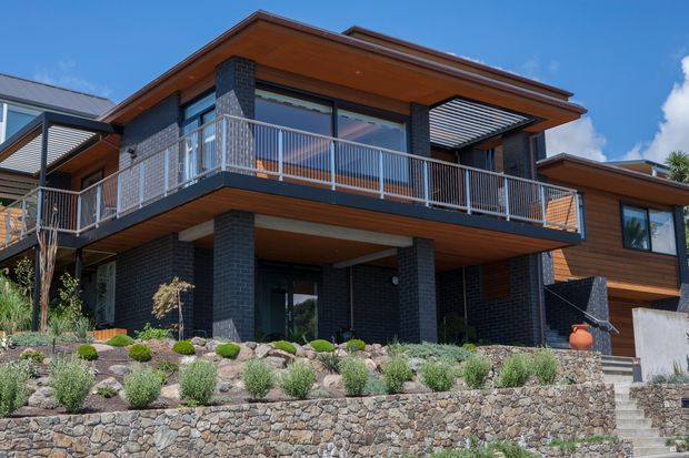 Exterior view of a contemporary multi-level home on a landscaped, sloping site. The house features dark brick masonry, extensive warm timber soffits and wall cladding, broad overhanging roof planes, large glazing, and an upper-level balcony with metal balustrades. Stone retaining walls, planted garden beds, and exterior steps define the foreground.