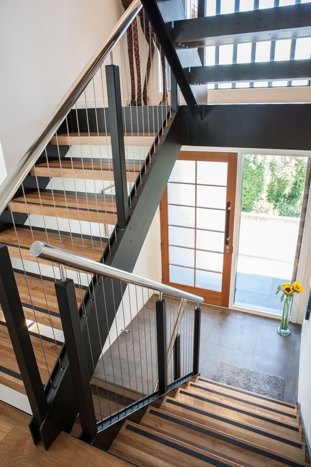 A bright, multi-level interior stairway viewed from above, featuring open-riser timber treads, black-painted steel stringers and posts, and slender vertical cable balustrades with stainless-steel handrails. The stair overlooks a tiled entryway with a timber-framed glazed front door and large windows, creating a strong double-height sense of space. Natural daylight enters through extensive glazing.