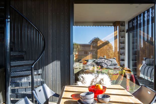 A dark, timber-lined dining area with a rectangular dining table and modern chairs sits beside a dramatic black spiral stair. Full-height glazing and a large sliding glass door open views into a bright bedroom, while built-in shelving frames the opening. The interior combines strong black vertical wall cladding with warm timber surfaces, abundant natural light, and colourful patterned bedding visible beyond.