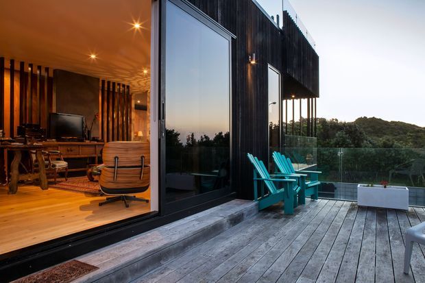 A contemporary indoor-outdoor living space with a dark timber-clad facade opening onto a spacious weathered timber deck. The interior visible through full-height glazing features warm timber flooring, a wood-burning fireplace or dark feature wall, timber furniture, a desk, and a lounge chair. Bright turquoise outdoor Adirondack chairs contrast with the charcoal exterior and muted natural landscape beyond. The scene has a relaxed, warm evening atmosphere and strong indoor-outdoor connection.