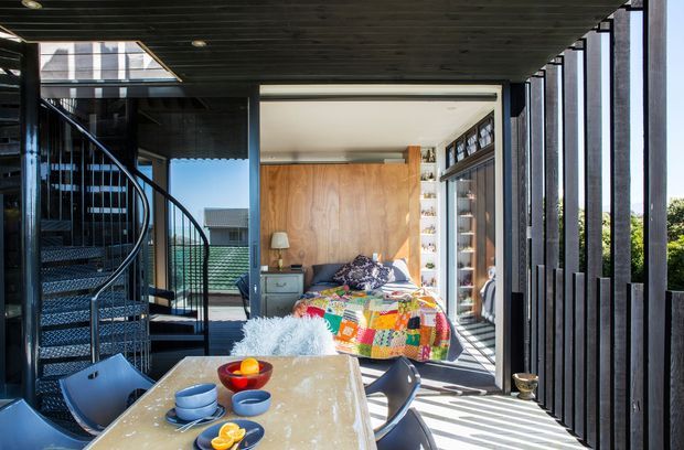 A bright contemporary open-plan interior with a dining table in the foreground, a bedroom visible beyond, and a sculptural spiral stair at left. Full-height glazing and dark vertical timber screens create strong filtered sunlight, while warm timber wall panelling and built-in shelving frame the sleeping area.