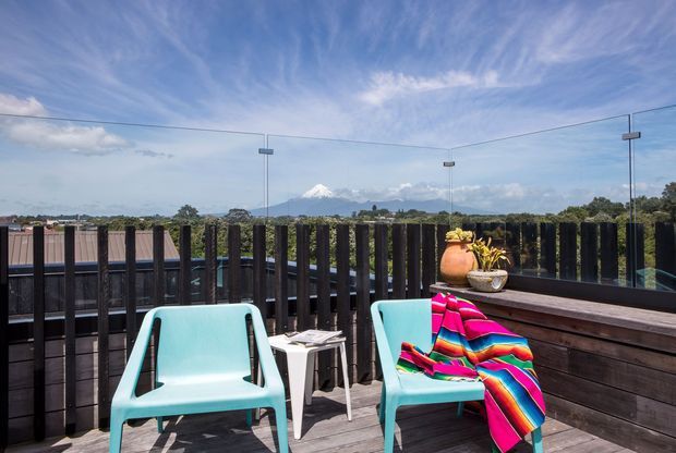 A sunny elevated outdoor deck or balcony with dark timber decking and privacy screening, clear glass balustrades, and two turquoise outdoor chairs around a small white side table. A colourful striped throw adds a vivid accent, while the view beyond includes trees, open landscape, and a distant snow-capped mountain under a blue sky.