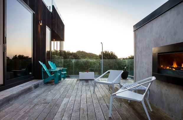 Contemporary outdoor terrace with weathered timber decking, clear glass balustrades, dark vertical timber-clad walls and a sculptural concrete outdoor fireplace. Teal Adirondack-style chairs and white lounge chairs are arranged for relaxation, with low vegetation visible beyond the terrace.