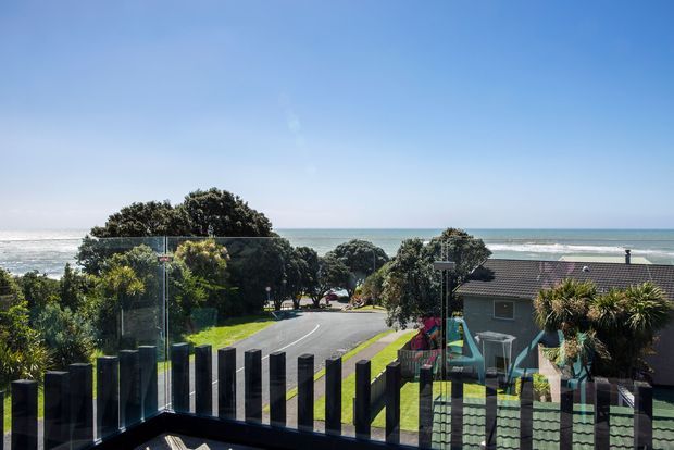 Elevated exterior view over a coastal residential neighbourhood, with the ocean and surf directly beyond the houses. A glass balustrade and dark vertical architectural screen frame the foreground terrace or balcony, while landscaped greenery and a curving residential road lead toward the beach.
