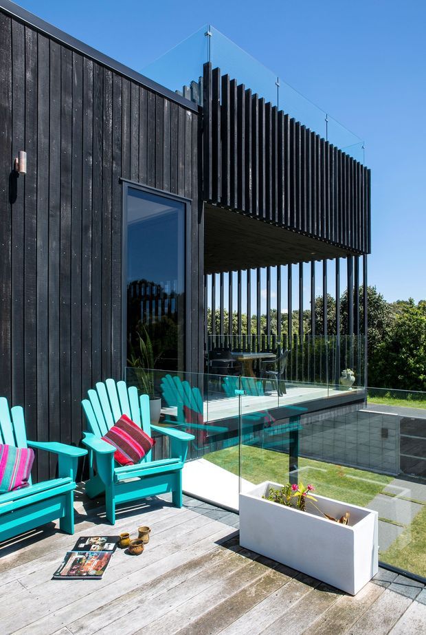 Exterior architectural photograph of a contemporary dark-clad house with a projecting upper volume and strong vertical timber fins. A glass balustrade encloses a timber deck with bright turquoise outdoor lounge chairs, while a lower paved terrace, lawn and planter are visible below. Clear blue sky and landscaped greenery surround the home.