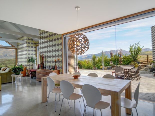 Bright open-plan dining area with a long light timber dining table and modern white chairs, polished concrete flooring, patterned feature wallpaper, and a sculptural spherical pendant light. Full-height glazing opens to a timber deck and landscaped outdoor entertaining area, with mountain scenery visible beyond.