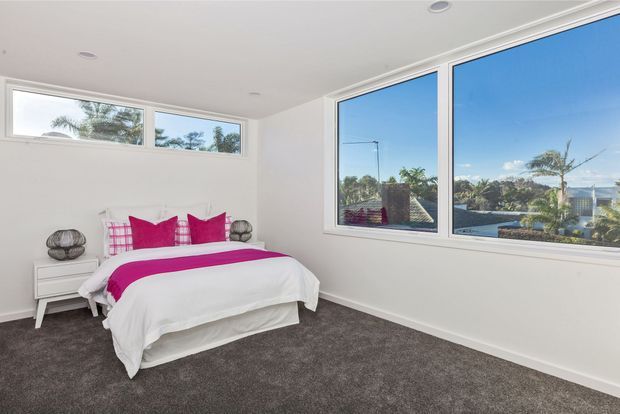 Bright, contemporary bedroom with a low-profile bed dressed in white bedding and vivid fuchsia cushions and throw. The room has white walls and ceiling, grey carpet, simple white bedside storage, and extensive windows offering leafy suburban views. The uncluttered fit-out creates an airy, clean atmosphere.