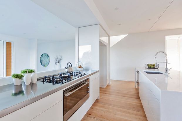 Bright contemporary open-plan kitchen with white handleless cabinetry, a long stone island with integrated gas cooktop and built-in oven, and a separate white island or benchtop with an undermount sink and spring tap. Light timber flooring, white walls and ceilings, extensive glazing, and minimal decorative styling create an airy, clean interior.