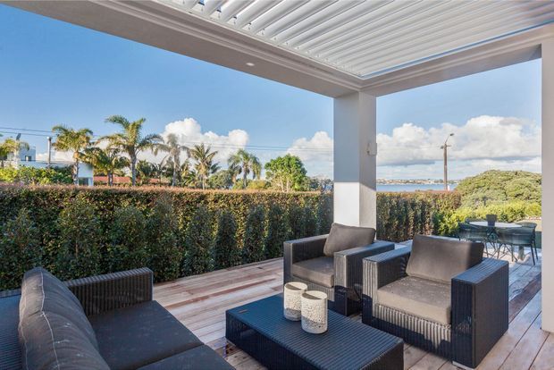 A covered outdoor terrace furnished as a relaxed lounge, with dark woven seating, a low coffee table, timber decking, and a white louvred roof structure. A hedge provides privacy, with palm trees, blue sky, and a distant water view beyond.