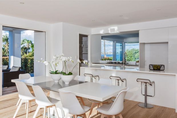 Bright, open-plan contemporary kitchen and dining area with a large white island, integrated sink and stainless tapware, bar seating, white dining table and sculptural white dining chairs. Floor-to-ceiling glazing opens to lush greenery and a water view, while light timber flooring and minimal white cabinetry create an airy, refined interior.