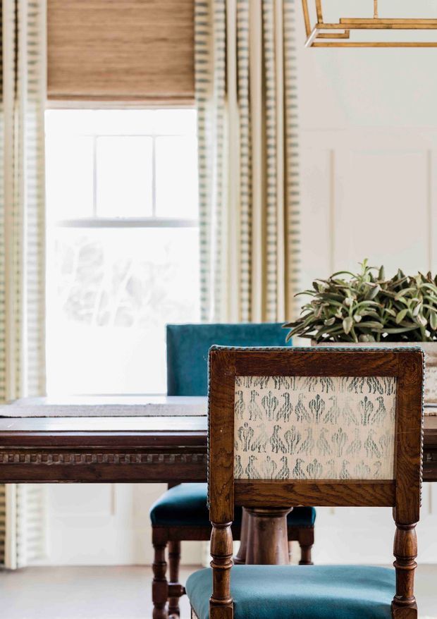 A close-up interior view of a refined dining area featuring a dark timber dining table and turned-leg upholstered dining chairs with teal-blue velvet seats and backs. A patterned fabric panel is visible on the foreground chair, with a potted plant on the table. The room is bright, with tall windows dressed in timber-toned blinds and patterned curtains, and a contemporary geometric pendant light overhead.