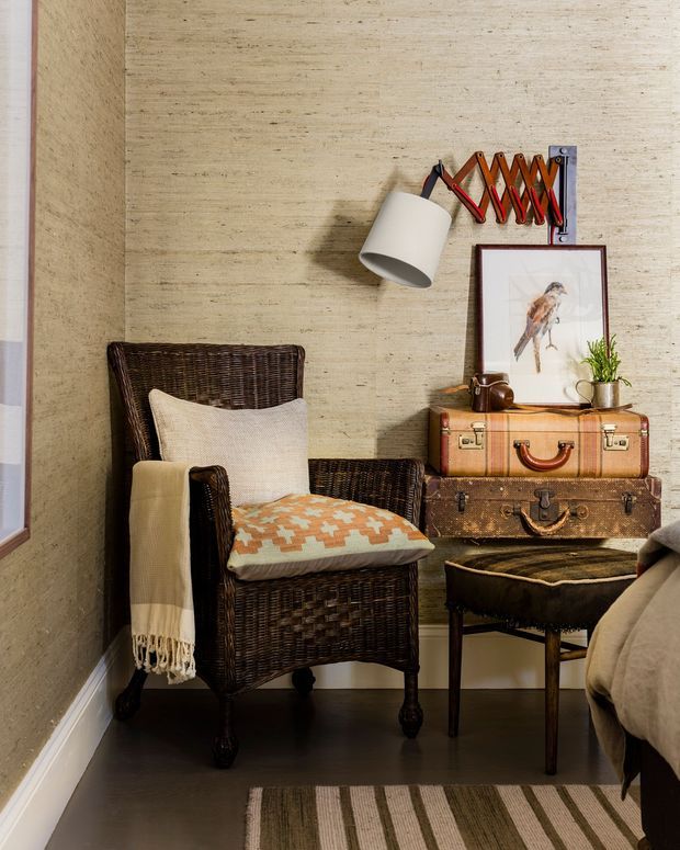 A warmly styled bedroom corner featuring a dark woven wicker armchair with cushions and a throw, a small upholstered bench, stacked vintage suitcases used as a side table, a framed bird print, potted plant, wall-mounted adjustable lamp, and a textured beige wall. The room has dark flooring, white trim, and a striped area rug, creating a cozy vintage-inspired reading nook.