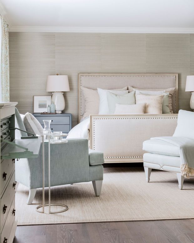 Bright, refined bedroom with a large upholstered bed featuring nailhead trim, layered neutral bedding, matching bedside lamps, a dark painted bedside chest, upholstered armchairs, a slim side table, and a pale area rug over timber flooring. The walls have subtle textured wallpaper with white decorative ceiling trim, creating a calm, traditional luxury interior.