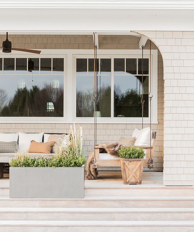 A bright covered outdoor porch or terrace with pale shingle-clad walls, large white-framed divided-light windows, a suspended wooden swing, outdoor seating, planters, and broad timber steps. The space has a relaxed coastal character with soft neutral furnishings and greenery.