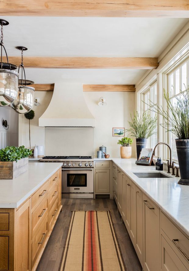 A bright, warmly styled galley kitchen with extensive pale wood and painted cabinetry, white countertops, a large farmhouse-style sink, stainless-steel range, and a sculptural plaster range hood. Exposed light timber ceiling beams, pendant lanterns, large divided windows, potted greenery, and a striped runner add character and texture.