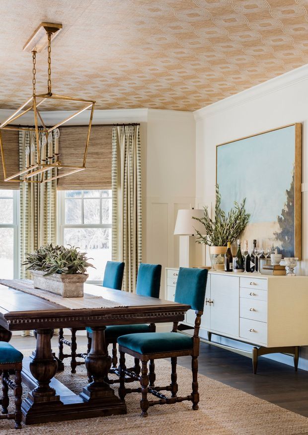 A refined dining room with a large dark timber trestle dining table, upholstered teal dining chairs, a pale sideboard, large artwork, layered window treatments, and a geometric brass pendant light. The room features a decorative patterned ceiling, traditional wall mouldings, dark timber flooring, and a neutral woven rug. The overall look blends classic architectural detailing with contemporary furnishings and vivid blue-green accents.