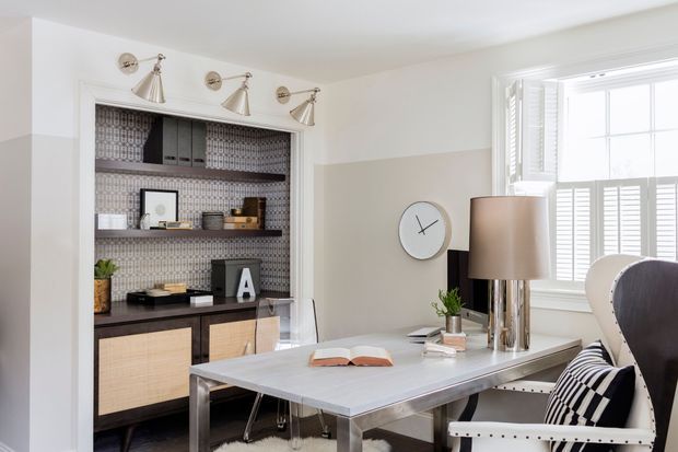 Bright, refined home office with a large desk positioned beside a shuttered window. The room features a built-in wall storage and display unit with patterned tile backing, dark shelving and benchtop, woven-front lower cabinetry, decorative lighting, a desk lamp, and a modern armchair.