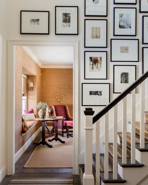 A refined traditional hallway and stairway with white painted trim, dark timber handrail, carpeted stairs, and a gallery wall of black-framed artwork. Through the open doorway is a compact sitting or reading room with warm wood wall panelling, built-in window seating, a small table, and red upholstered chairs.