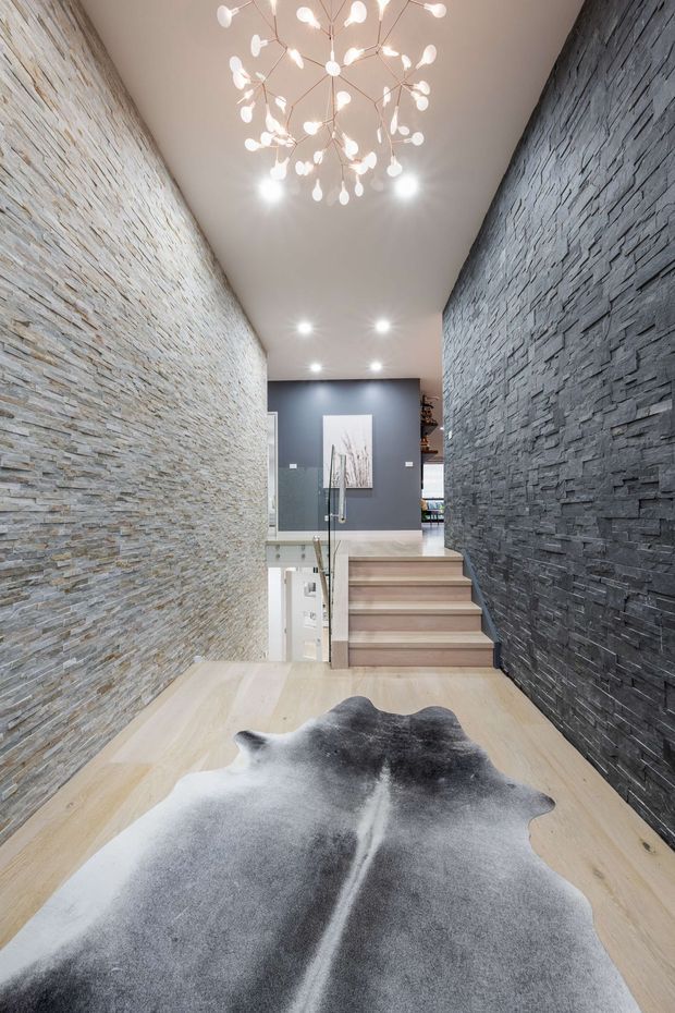 A tall, narrow interior stairway and landing with dramatic stacked-stone feature walls on both sides. Pale timber flooring leads to a short flight of timber stairs with a glass balustrade, while a sculptural branching pendant light hangs from the smooth white ceiling. A dark grey wall with framed artwork is visible at the upper landing, and a monochrome cowhide rug occupies the foreground.