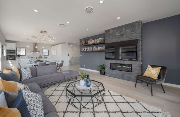 Open-plan contemporary living room with a large grey sectional sofa, geometric patterned rug, black metal coffee table, and a dark feature wall incorporating a grey stone-clad fireplace and wall-mounted television. Built-in open shelving displays objects beside the fireplace. The kitchen and dining area are visible in the background, with light timber flooring, white cabinetry, pendant lights, and a decorative chandelier.